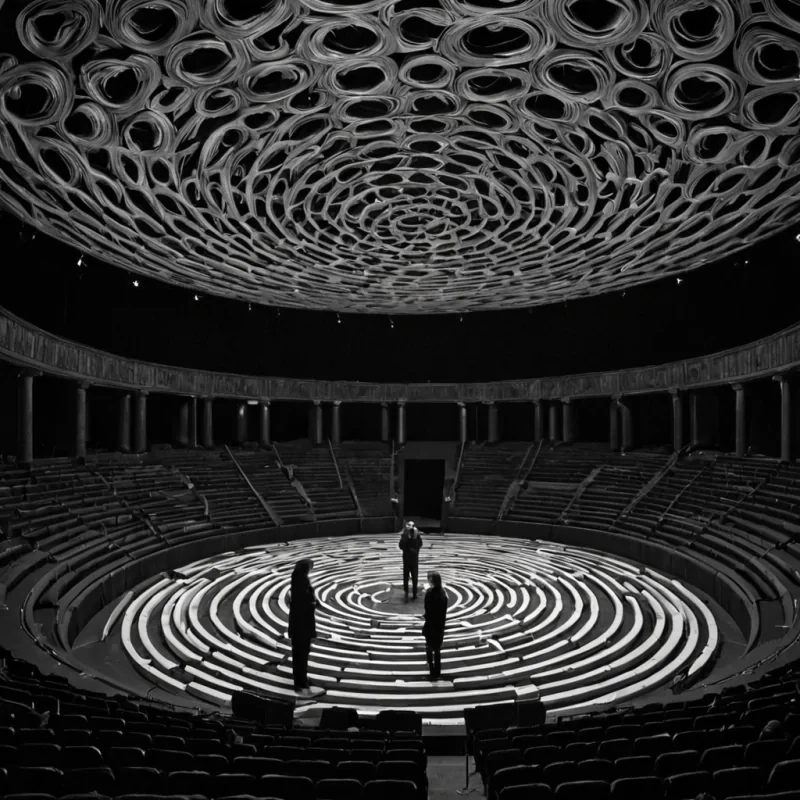 A single, selffragmenting thread of reversed entropy, shaped like a spiraling, threedimensional lattice of unformed regret, drifts through the center of a vast, inverted theater constructed from the compressed, unperformed scenes of a civilization that communicated through the duration of synchronized, silent rehearsing of the same unspoken monologue. The thread does not unravelit unregrets, each pulse dissolving a layer of emotional weight that never coalesced into a shared performance, reforming into ephemeral, amberveined afterimages shaped like the negative space between two mouths that never opened in unison in the same moment of mutual silence. The theater is not dramaticit is a stratified expanse of petrified anticipation, each